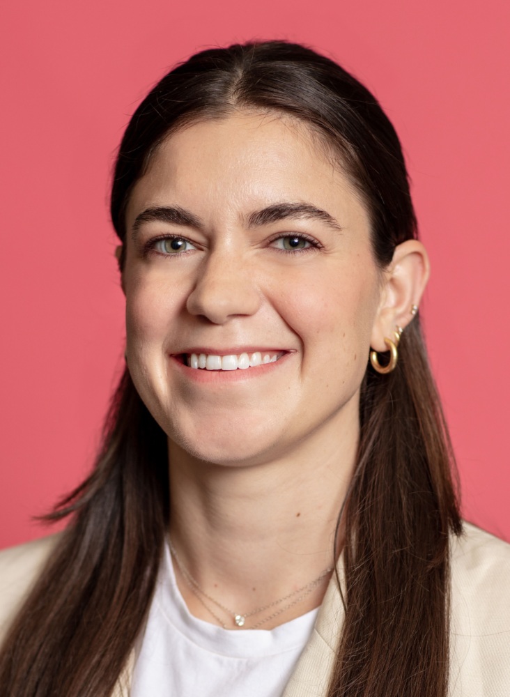 A woman with long, dark brown hair smiles at the camera. She is wearing a light-colored blazer, a white shirt, and gold hoop earrings, with a pink background behind her.